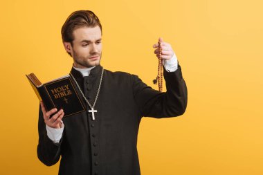 thoughtful catholic priest holding bible and looking at wooden rosary beads isolated on yellow