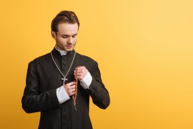 young thoughtful catholic priest holding wooden rosary beads isolated on yellow
