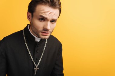 young thoughtful catholic priest looking at camera isolated on yellow clipart