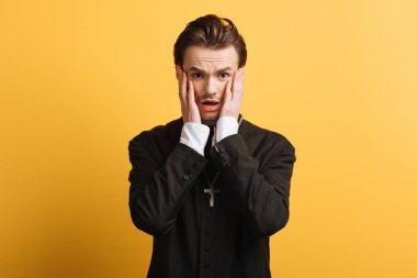 shocked catholic priest touching face while looking at camera isolated on yellow