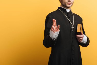partial view of catholic priest showing blessing gesture while holding bible isolated on yellow