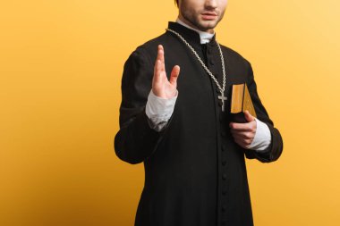 cropped view of catholic priest showing blessing gesture while holding bible isolated on yellow