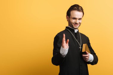 smiling catholic priest showing blessing gesture while holding bible isolated on yellow