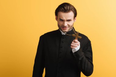 angry catholic priest showing cross while looking at camera isolated on yellow clipart