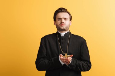 young catholic priest with bored face expression holding cross and looking at camera isolated on yellow clipart