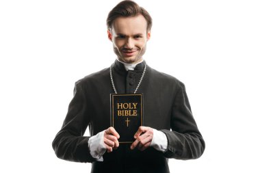 young, smiling catholic priest showing holy bible isolated on white