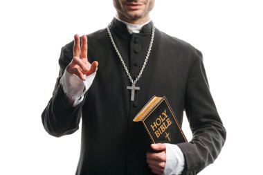 cropped view of young catholic priest holding holy bible and showing blessing gesture isolated on white