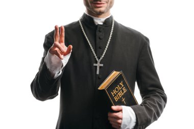 cropped view of young catholic priest holding holy bible and showing blessing gesture isolated on white clipart
