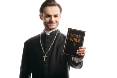 young catholic priest smiling while holding holy bible isolated on white