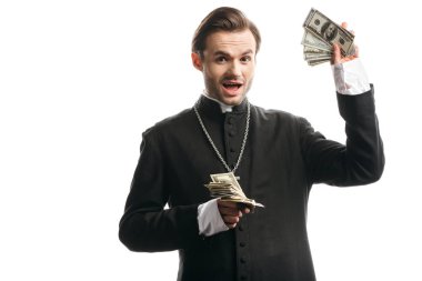 excited catholic priest looking at camera while showing dollar banknotes isolated on white