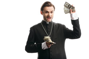 excited catholic priest looking at camera while showing dollar banknotes isolated on white clipart
