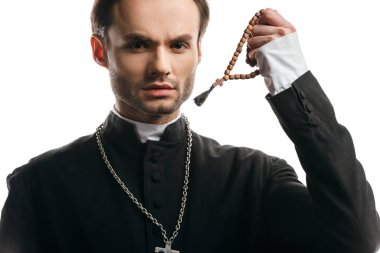young, serious catholic priest holding wooden rosary beads while looking at camera isolated on white
