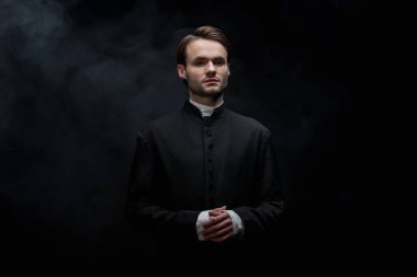 young serious catholic priest standing with folded hands on black background with smoke