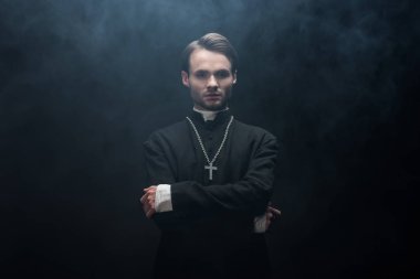 young serious catholic priest standing with crossed arms on black background with smoke