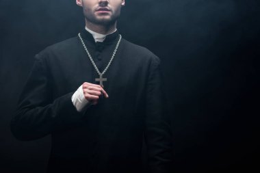 cropped view of catholic priest touching silver cross on his necklace on black background with smoke clipart