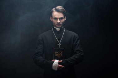 young confident catholic priest looking at camera while holding holy bible on black background with smoke