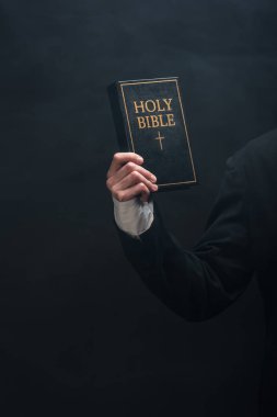 partial view of catholic priest holding holy bible isolated on black clipart