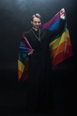 Elinde lgbt bayrağı olan gülümseyen Katolik rahibin siyah arka plandaki dumanlı kameraya tam boy görüntüsü