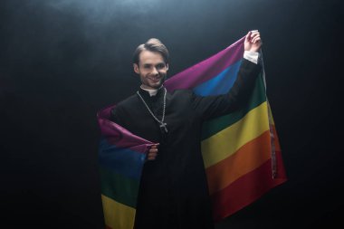 Elinde lgbt bayrağı tutan gülümseyen Katolik rahip siyah arkaplanda dumanla kameraya bakıyor.