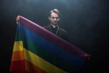 Elinde lgbt bayrağı tutan gülümseyen Katolik rahip siyah arkaplanda dumanla kameraya bakıyor.