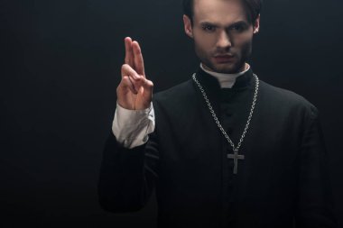 young confident catholic priest looking at camera and showing blessing gesture isolated on black clipart