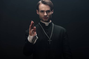 young confident catholic priest looking at camera and showing blessing gesture isolated on black clipart