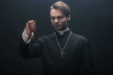 young tense catholic priest looking at wooden rosary beads isolated on black clipart
