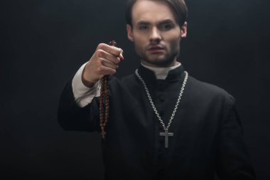 Genç kendinden emin katolik rahibin seçici odak noktası, siyah üzerine izole edilmiş, uzanmış ellerde ormanlık tespih boncukları tutan.