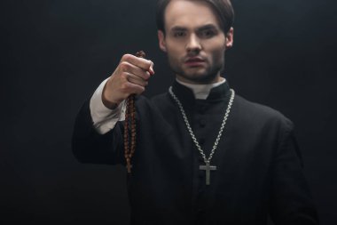 selective focus of young confident catholic priest holding wooded rosary beads in outstretched hand isolated on black clipart