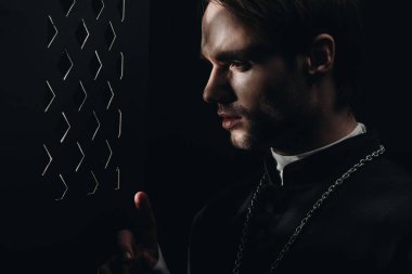 young tense catholic priest looking through confessional grille in dark with rays of light clipart
