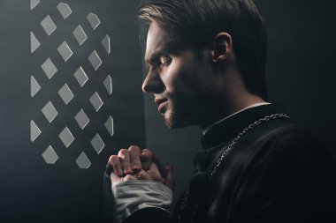 young thoughtful catholic priest praying with closed eyes in dark near confessional grille with rays of light clipart