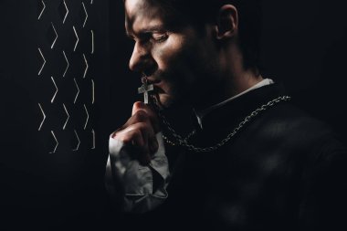 young tense catholic priest kissing cross on his necklace in dark near confessional grille with rays of light clipart