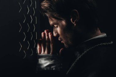 young catholic priest praying with closed eyes near confessional grille in dark with rays of light clipart