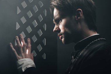 young tense catholic priest looking through confessional grille in dark with rays of light clipart