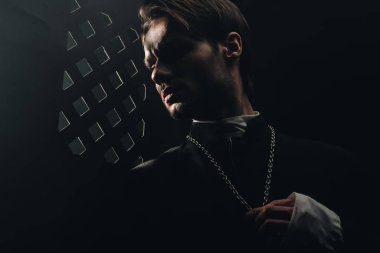 young serious catholic priest touching cross on his necklace in dark near confessional grille clipart