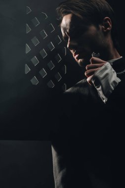 young serious catholic priest touching cross on his necklace in dark near confessional grille clipart