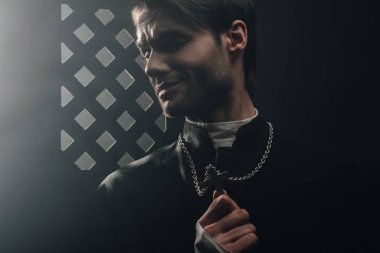 young worried catholic priest touching cross on his necklace in dark near confessional grille clipart