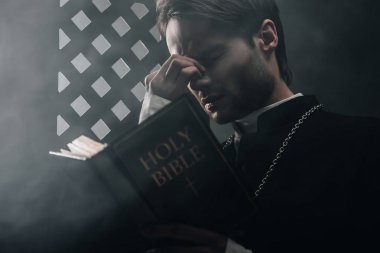 young thoughtful catholic priest touching face while reading bible near confessional grille in dark with rays of light clipart