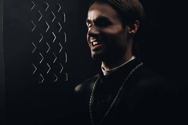 young sarcastic catholic priest laughing near confessional grille in dark with rays of light