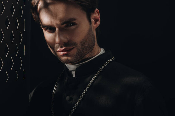 young confident catholic priest looking at camera near confessional grille in dark with rays of light