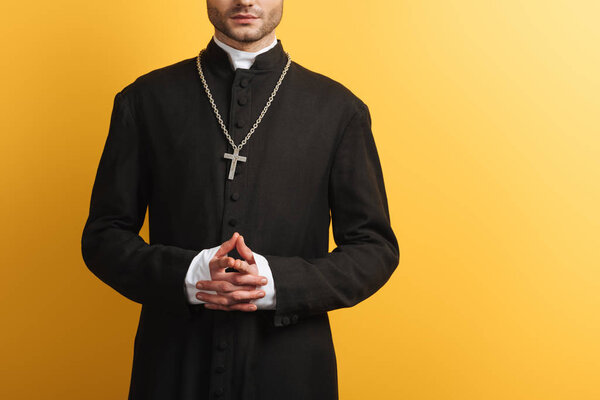 cropped view of catholic priest standing with clenched hands isolated on yellow