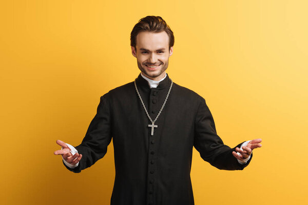 smiling catholic priest standing with open arms and smiling isolated on yellow