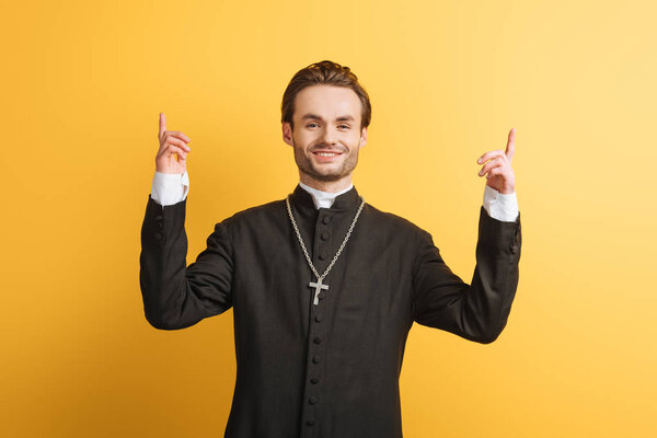 smiling catholic priest pointing up with fingers while looking at camera isolated on yellow