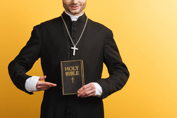 cropped view of catholic priest pointing with finger at holy bible isolated on yellow