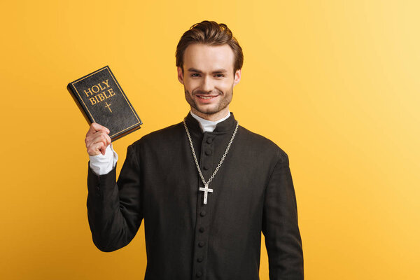 happy catholic priest holding bible while smiling at camera isolated on yellow