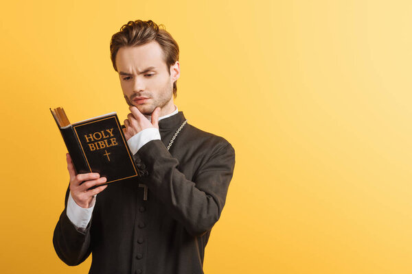 concentrated catholic priest touching face while reading bible isolated on yellow