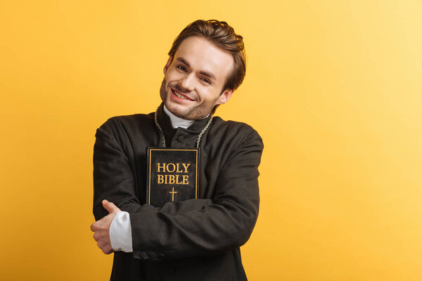 happy catholic priest looking at camera while hugging bible isolated on yellow