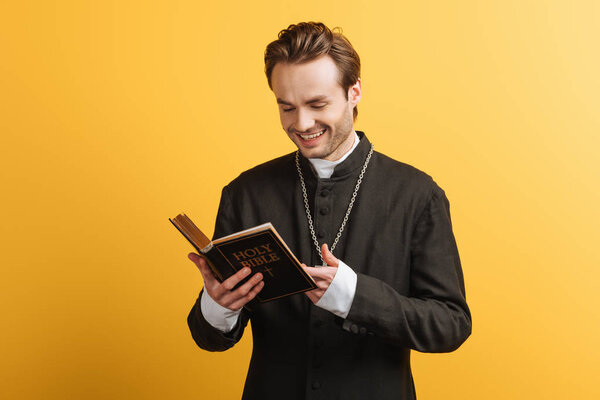 cheerful catholic priest laughing while reading bible isolated on yellow
