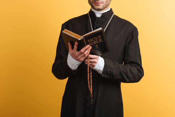 cropped view of catholic priest reading bible and holding wooden rosary beads isolated on yellow