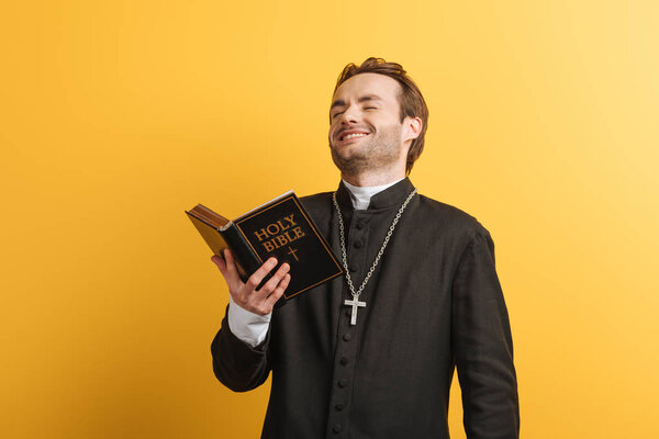 excited catholic priest laughing while holding holy bible isolated on yellow